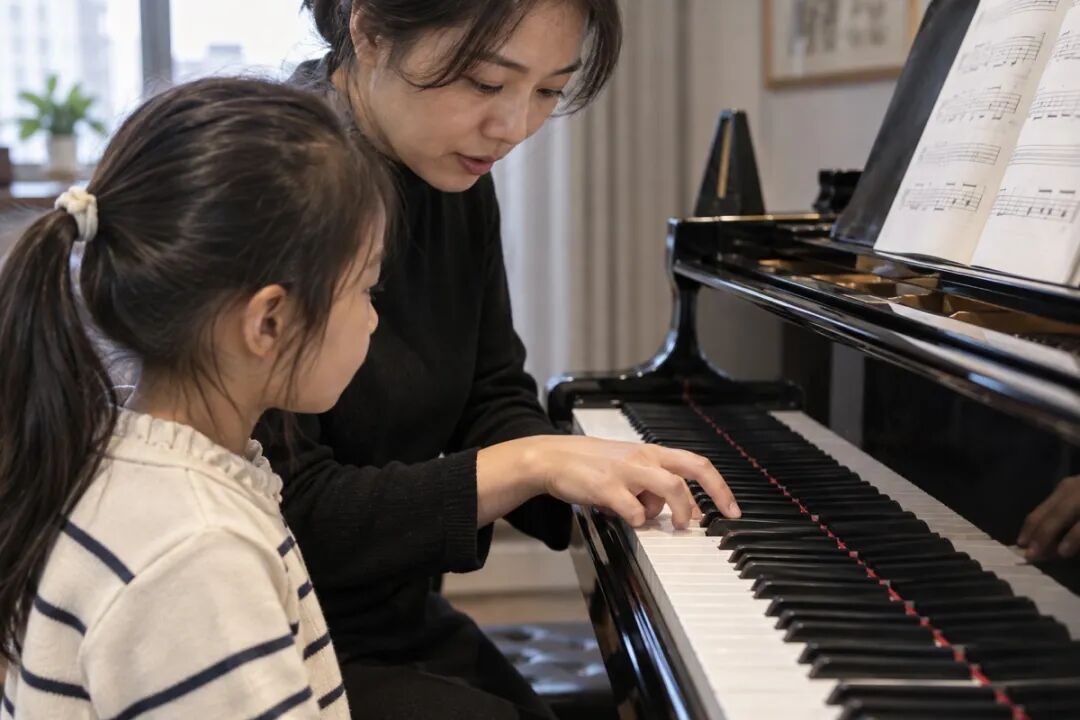 A trial lesson seen from a parent's perspective: the teacher demonstrates just two notes while the child stands beside them, watching the hands carefully; the view focuses on the hands and keyboard, capturing a natural teaching moment.