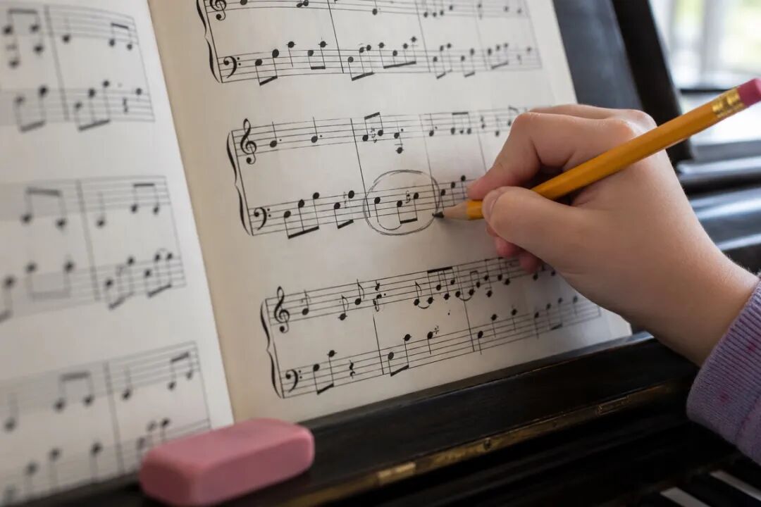 A child's hand holds a yellow pencil and circles a bar on the bass staff of a piano score, showing how children can mark the passage their teacher pointed out after a lesson.